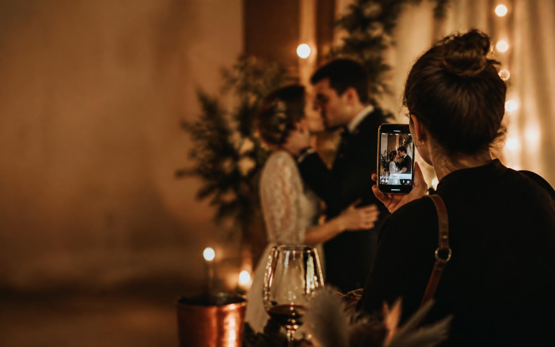 Storymaker de costas filmando com o celular o beijo de um casal em um casamento elegante